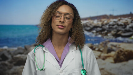 Woman doctor with curly hair holding a green stethoscope against her white coat at a rocky seaside;...
