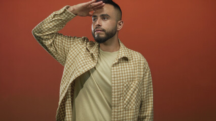 Man shielding eyes with hand in studio against orange backdrop wearing checked shirt and tee, looking into distance; curiosity exploration.