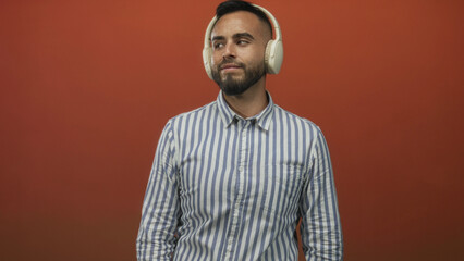 Young hispanic man wearing white headphones and striped shirt shrugs in studio against orange wall; playful.