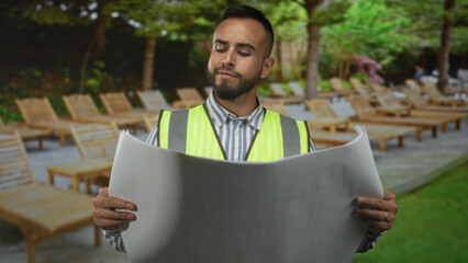 Man engineer holding large rolled blueprint in park near lounge chairs wearing reflective vest; calm planning focus.