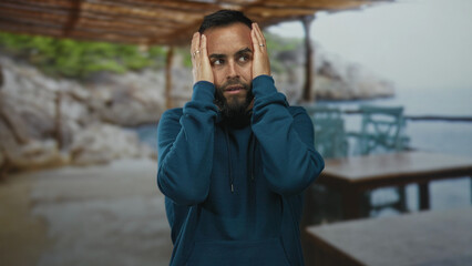 Young hispanic man in blue hoodie with hands on cheeks at beach under wooden pergola by sea; contemplation.