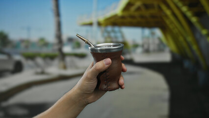 Person holding traditional mate cup with straw on urban street background, showcasing outdoor city setting and cultural beverage.