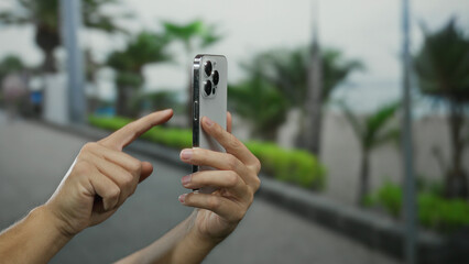 Man using smartphone outdoors in a city street, showcasing modern technology and digital communication in an urban environment with blurred background trees.