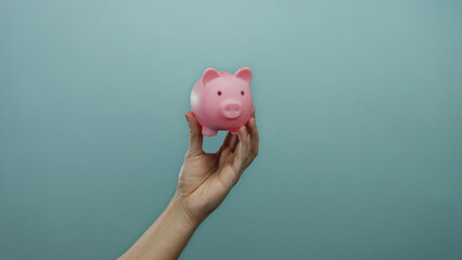 Hand of man holding pink piggy bank against blue wall background showcasing savings concept in an...