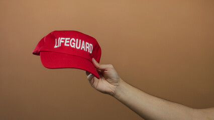 Caucasian man holding red lifeguard cap against brown isolated background wall.