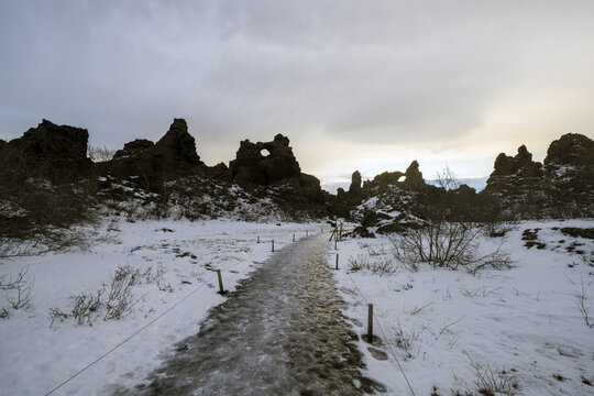 View of a path cutting through a snow-covered landscape, flanked by jagged, dark rock formations under a cloudy sky, Blonduos, Iceland.