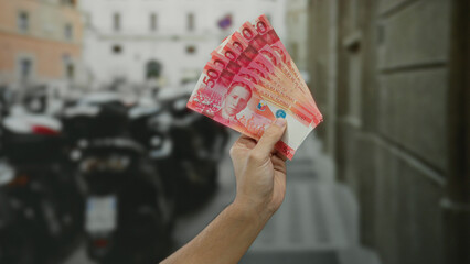 Man holding philippine pesos on city street with motorcycles in background suggesting economic...