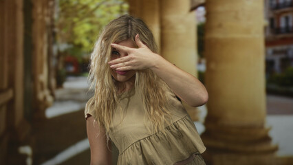 Woman covers face with hand and peeks through fingers beside weathered columns on an old town street; shyness curiosity.