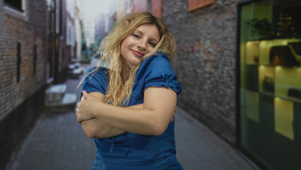Naklejka premium Woman hugging her arms on a narrow street alley wearing blue dress, bare forearms visible; self care contentment.