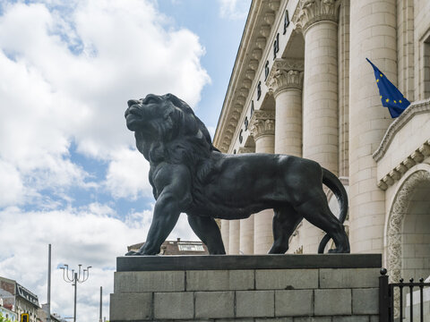 View of a lion statue standing proudly against the backdrop of a grand building with tall columns and a blue European Union flag, Sofia, Sofia City Province, Bulgaria.