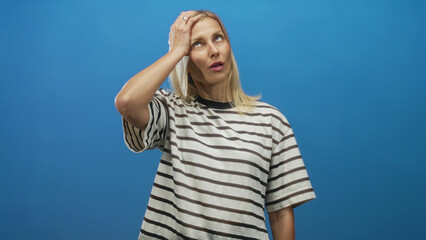 Woman touching temple and side of face with hand in studio against blue backdrop, striped tee, pensive expression and upward glance; contemplation.