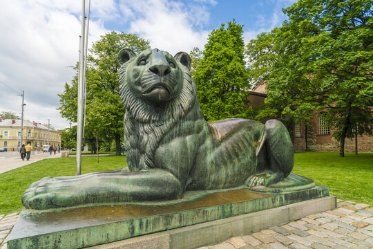 View of a majestic verdigris lion statue rests regally on a stone plinth amidst vibrant green grass, with soft blue skies above, Sofia, Sofia City Province, Bulgaria.