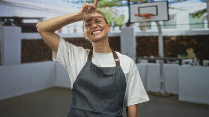 Woman in denim apron raises arm with hand to forehead on outdoor basketball court near hoop and white wall  cheerful. © Krakenimages.com
