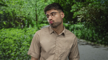 Hispanic young man with glasses puckers lips on forest path amid green foliage near shaded area; flirtation.
