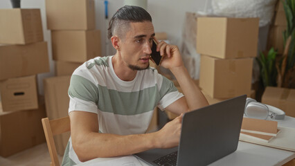 Man holding phone to ear and using laptop amid stacked moving boxes in a building; relocation stress.