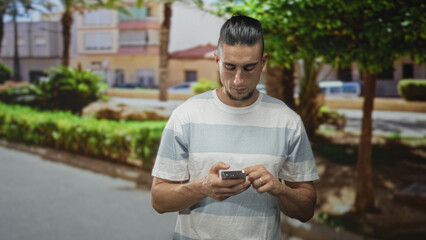 Obraz premium Man tapping smartphone with both hands and looking down on a sunny street with trees and buildings; focused connectivity.