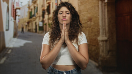 Fototapeta premium Woman pressing hands together in prayer with closed eyes on a narrow old town street; solemn prayer.