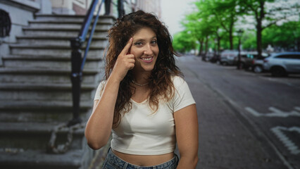 Woman points finger to temple while smiling, wearing a white crop top and jeans on a city street;...