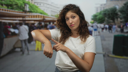 Woman points finger to wrist at outdoor city market street with visible forearm and hand gesture among souvenir stalls; time reminder impatience.
