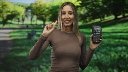 Woman holds a card reader and raises index finger while standing in a forest with green foliage and grass; confidence payment trust.