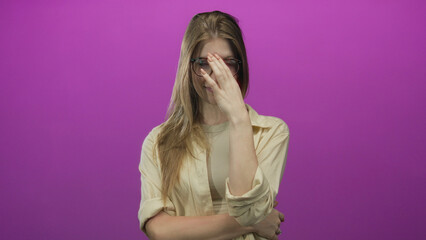 Woman blonde adjusts glasses with hand touching forehead and crosses arm in pink studio; embarrassment.