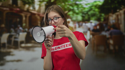 Young blonde woman volunteer in red shirt holding megaphone and pointing finger on street terrace; empowerment.