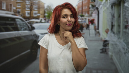 Woman with hand on chin touching red hair in white t shirt on street; thoughtful pondering curiosity.