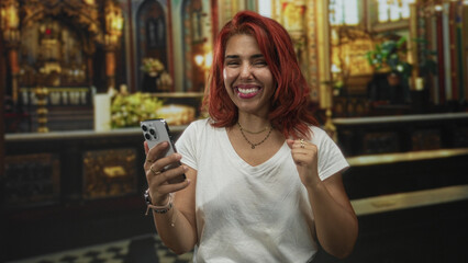 Young hispanic woman smiling and holding a smartphone in a fist pump gesture in an ornate catholic church; celebration joy hope.