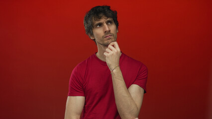Man wearing red t shirt holds hand to chin gesture in a red studio scene for thoughtful expression; thinking.