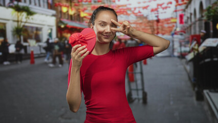 Young woman holds red heart shaped giftbox and flashes peace sign fingers in busy street market; playful celebration.