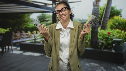 Young hispanic woman holding smartphone and fanned cash, shrugging shoulders while wearing glasses and olive blazer in building poolside patio with palm trees; indifference.