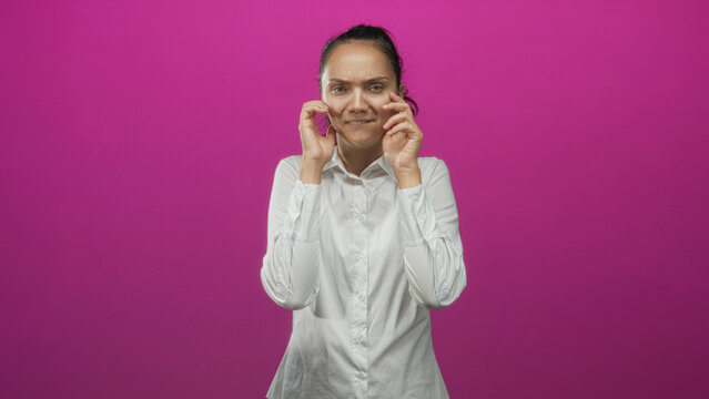 Woman in white shirt holds hands beside cheeks with splayed fingers and pursed mouth in studio with vivid pink backdrop; mild annoyance.