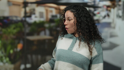 Fototapeta premium Woman standing on restaurant terrace, wearing glasses and striped sweater, with curly hair against a bustling outdoor street background, expressing surprise or curiosity.