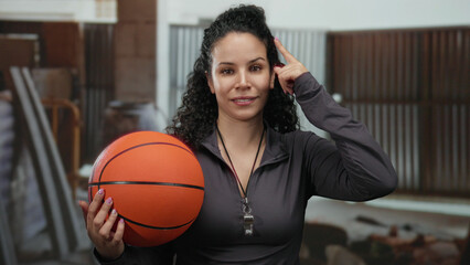 Woman holding basketball indoors with a whistle in a casual setting, wearing a black outfit, smiling, hispanic, thinking, curly hair, indoors, at workplace.