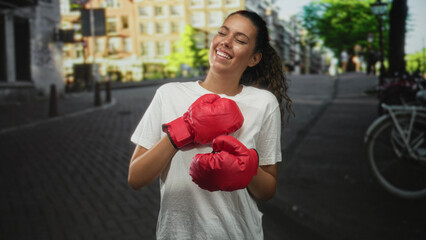 Hispanic teenage woman in white t shirt with red boxing gloves holds fists to chest on a cobblestone city street; joyful confidence.
