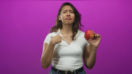 Young hispanic woman holding red apple and pointing to chest with puzzled expression in purple studio; doubt healthy choice.