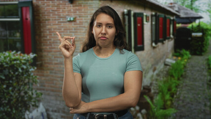 Woman pinches fingers and crosses arm at building entrance near window shutters in casual tshirt and jeans; mild frustration.