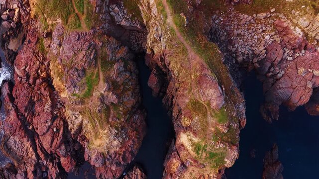 Aerial View of Rocky Cliffs with Path and Sea. Aerial shot of dramatic rocky cliffs along the coastline. Red and brown rock formations with patches of greenery and a narrow coastal path.