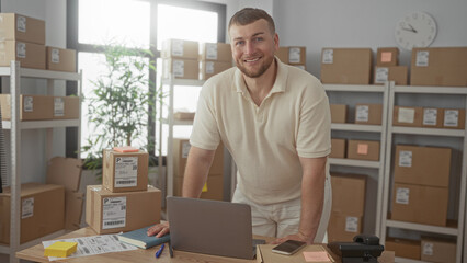Man leaning on desk with parcel boxes, laptop and smartphone in a building office, smiling at...