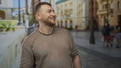 Fototapeta premium Man smiling and looking sideways on a city street in a short sleeved brown t shirt, beard visible; contentment curiosity travel.