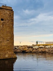 Woman gazing at the sea near Paphos Castle, enjoying the scenic coastal view with medieval stone walls in the background and a serene Mediterranean atmosphere