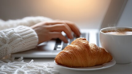 Individual is focused on typing on a laptop while enjoying a croissant and coffee in a welcoming indoor environment