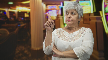 Woman points finger upward with crossed arm beside slot machines in a casino building, grey hair and white lace top, stern expression and raised eyebrow; firm disapproval.