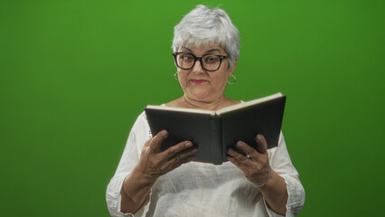 Senior woman with grey hair and glasses holding a hardcover book with hands, turning pages and smiling in a studio green screen; serenity reading.