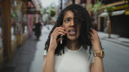 African american woman speaking angrily on smartphone on busy city street with storefronts and blurred passersby; frustration.