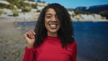 Woman dancing with raised hands and a smiling face on a sandy beach shoreline under a clear blue sky in a red top; joyful freedom.