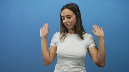 Fototapeta premium Young woman raises hands palms outward in a blue studio, shrugging in a white tshirt and looking away; indifference.