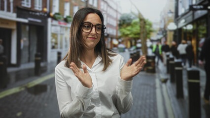 Woman clapping hands on a crowded urban street wearing white shirt and glasses with a wry smile and relaxed posture; approval encouragement.