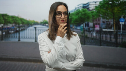Fototapeta premium Woman in white shirt touches chin while holding glasses frame, slight smile and folded arms on a street bridge by a canal in a city urban scene; thoughtful reflection.