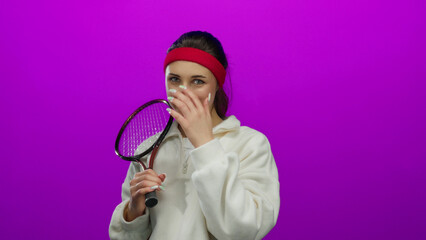 Woman posing with tennis racket on a vibrant pink background, exuding youthful energy and showcasing her athletic attire and playful demeanor while touching her cheek.